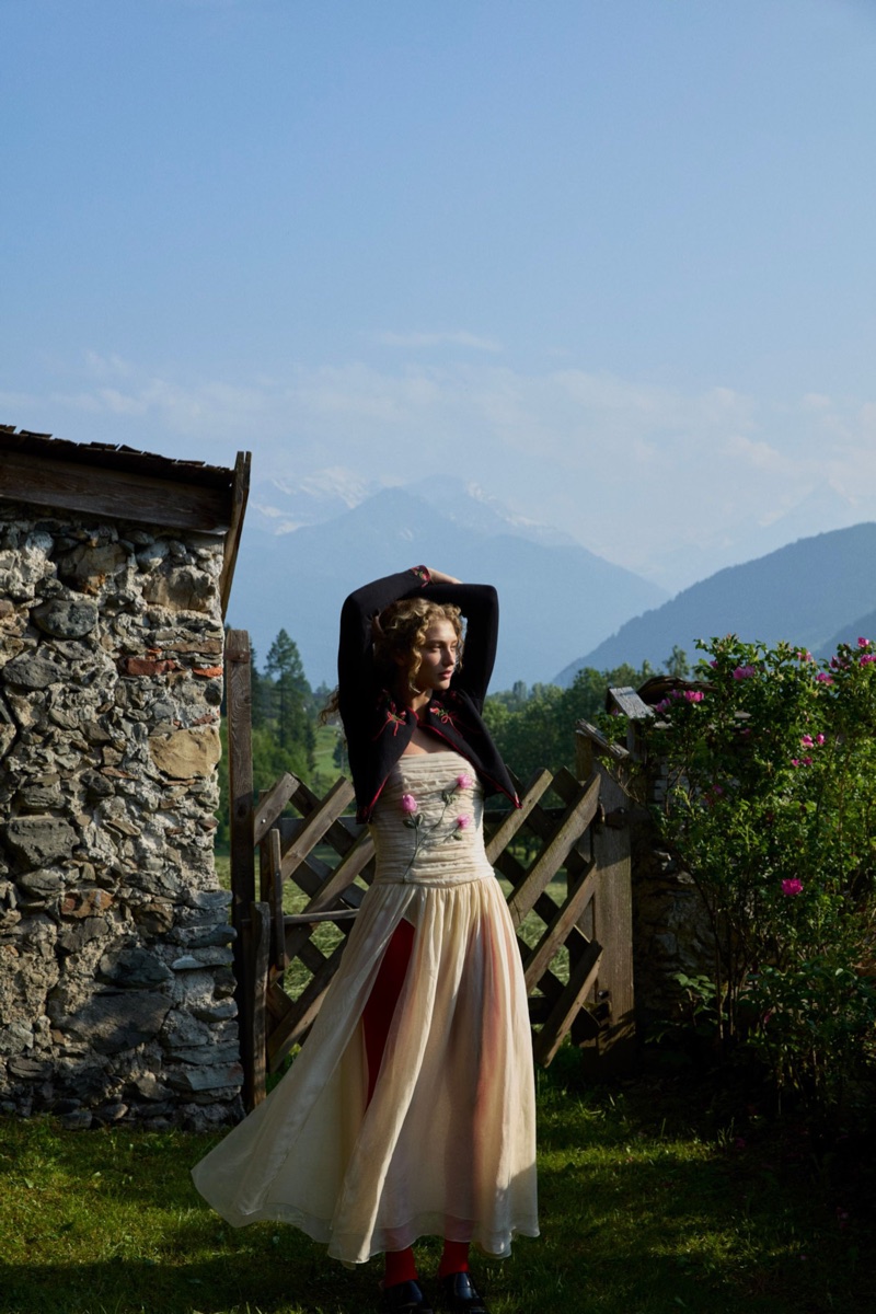 Model Dorit Revelis in a cream dress and dark jacket posing outdoors near a rustic stone wall and wooden gate under mountains.