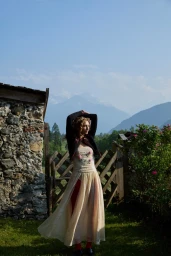 Model Dorit Revelis in a cream dress and dark jacket posing outdoors near a rustic stone wall and wooden gate under mountains.