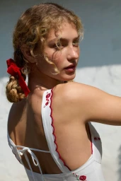 Model with blonde curly hair in twin braids tied with a red ribbon wearing a white dress with red floral embroidery