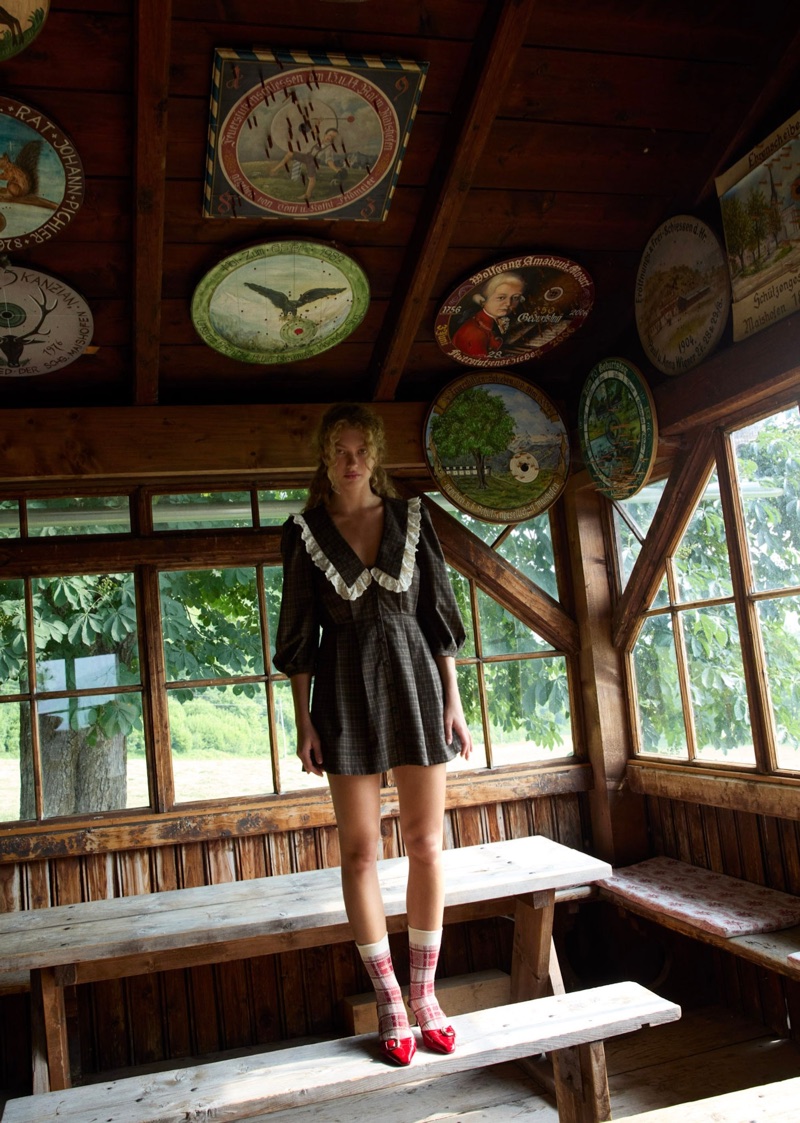 Model Dorit Revelis in a dark ruffled minidress standing on a rustic wooden bench inside a wood-paneled cabin decorated with painted plates.