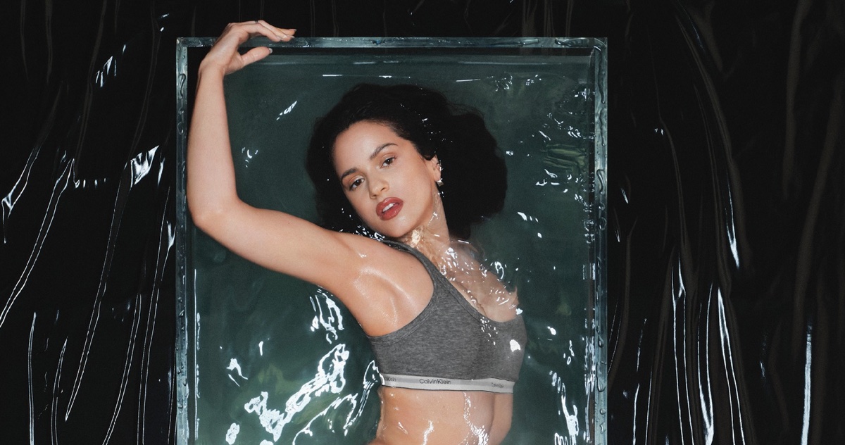 Rosalía posing underwater in a grey Calvin Klein sports bra within a rectangular glass tank against a crinkled black vinyl background.