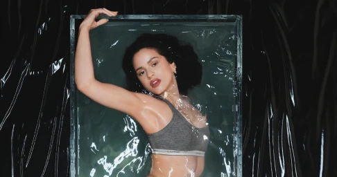 Rosalía posing underwater in a grey Calvin Klein sports bra within a rectangular glass tank against a crinkled black vinyl background.