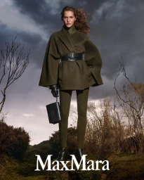 Model Angelina Kendall in olive green cape coat and leggings holding a black handbag under a stormy sky for Max Mara Fall-Winter 2025.