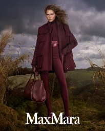Model Angelina Kendall in a burgundy coat and matching handbag outdoors against an overcast countryside landscape for Max Mara.