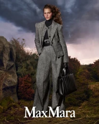 Model Angelina Kendall in a grey tweed suit and holding a black leather handbag outdoors under a dramatic stormy sky.