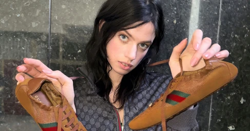 Model Lily McMenamy holding two brown sneakers against a dark stone wall for Gucci Shift, photographed by Tess Petronio.
