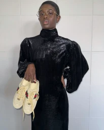 A model wearing a black turtleneck dress and large glasses holding a pair of white sneakers for Gucci Shift.