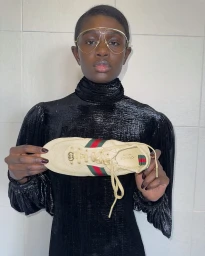 Model wearing oversized gold aviator glasses and black textured top holding a cream sneaker for Gucci Shift