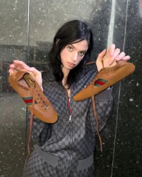 Model holding up a pair of brown sneakers in a campaign shot by Tess Petronio for Gucci Shift