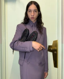 Model wearing a lavender suit jacket holding black sneakers in front of a pale green door for Gucci Shift.