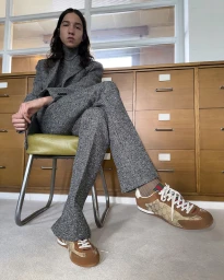 Model seated on a green-cushioned chair wearing a grey tweed suit and brown monogram sneakers.