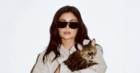 Kylie Jenner wearing black sunglasses and a beige jacket holding a striped cat against a white background