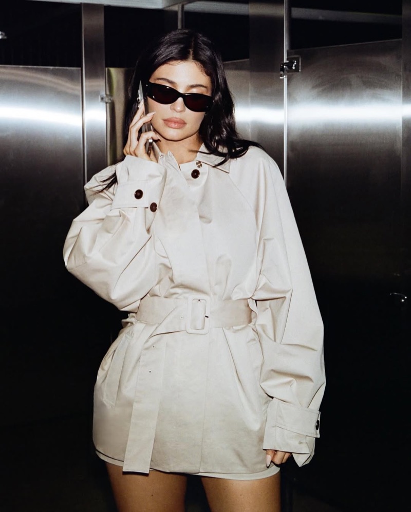 Kylie Jenner wearing black sunglasses and a beige belted trench coat while holding a phone in a metallic interior setting.