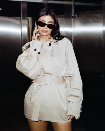 Kylie Jenner wearing black sunglasses and a beige belted trench coat while holding a phone in a metallic interior setting.