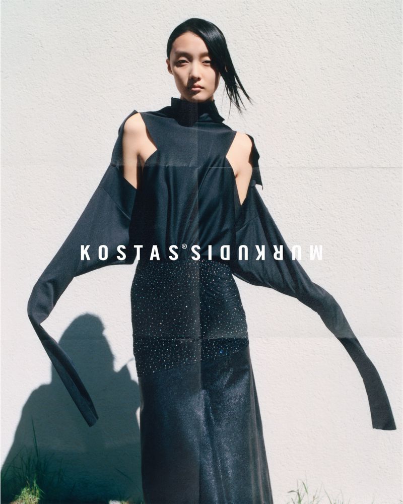 A model poses in an open-shoulder black dress against a white wall for a fashion advertisement, illuminated by bright, natural lighting.