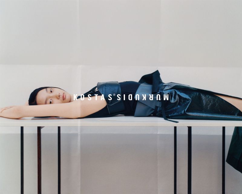 Fashion shot of a reclining adult female wearing a dark top and dark shiny skirt on a white table in front of a paper backdrop.