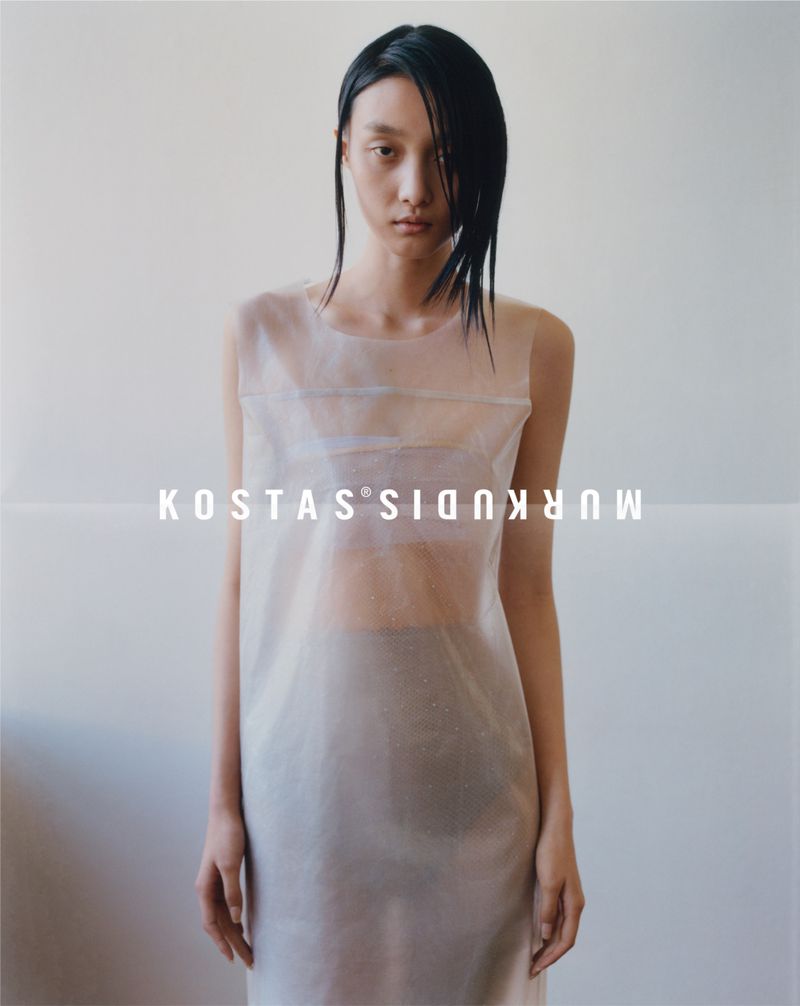 Portrait of a slender adult female fashion model with dark hair wearing a semi-sheer light grey sleeveless dress in a bright indoor setting.