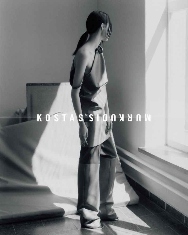 Fashion editorial in black and white features a slender Asian woman in stylish asymmetrical clothing near a bright window.