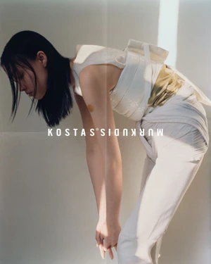 Model bent forward wearing white layered top and trousers against a neutral background with soft sunlight.
