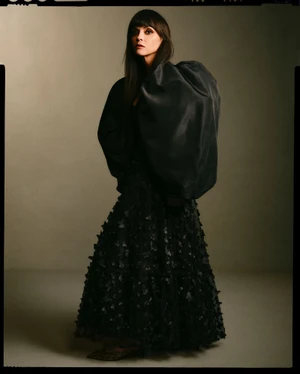 Christina Ricci wearing a voluminous black puff-sleeve top and textured skirt against a neutral studio backdrop.