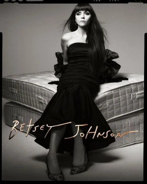 Christina Ricci sitting on stacked mattresses wearing a black strapless dress for the Betsey Johnson Fall 2025 campaign.