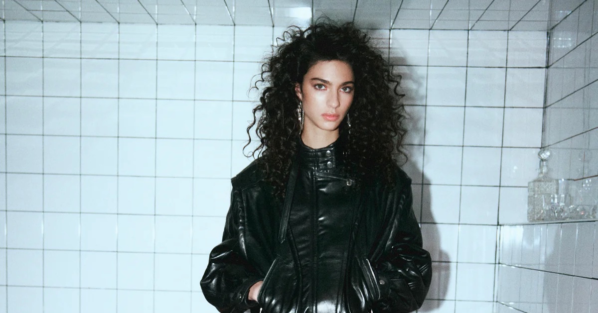 Model Loli Bahia with voluminous curly hair wearing an oversized black leather jacket against a white tiled wall.
