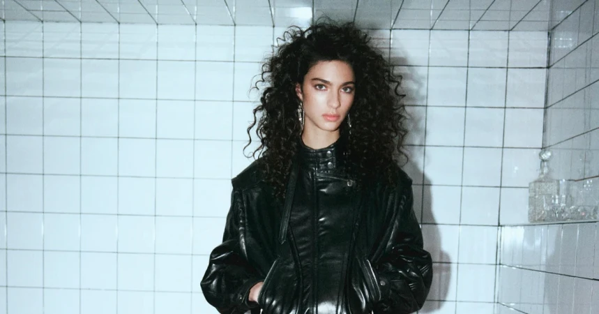 Model Loli Bahia poses in an oversized black leather jacket with voluminous curly hair against a white tiled wall.