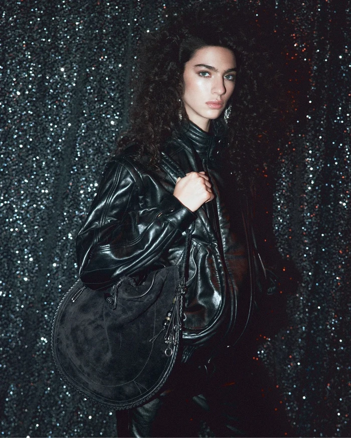 Model Loli Bahia in a black leather jacket holding a suede handbag against a glittering dark backdrop for Isabel Marant