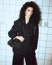 Model Loli Bahia with voluminous curly hair wearing a black pinstripe suit and holding a leather handbag against a tiled wall