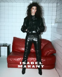 Model Loli Bahia in a black leather jacket and matching pants standing in front of a red sofa against tiled walls.