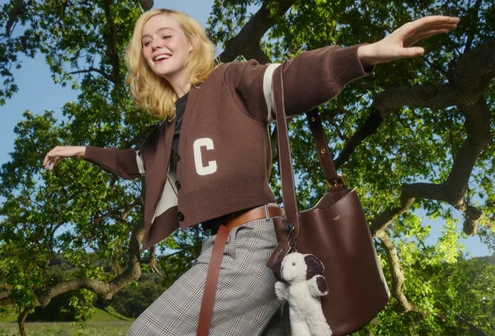 Elle Fanning balancing on a tree branch outdoors wearing a brown cardigan and plaid trousers with a leather bucket bag.