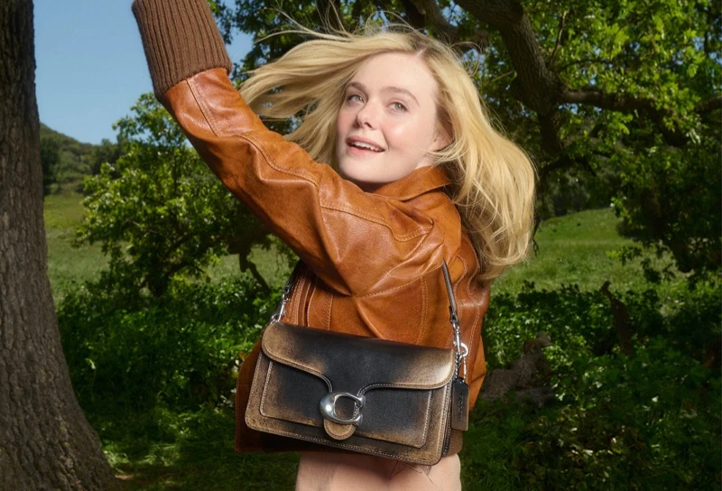 Elle Fanning wearing a brown leather jacket holding a black shoulder bag outdoors with trees in the background