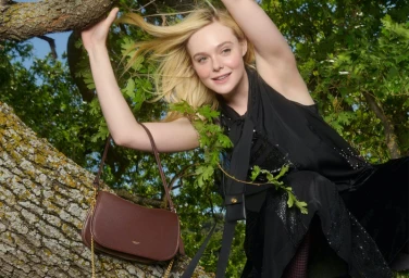 Elle Fanning holding up her blonde hair while posing in a tree branch next to a brown leather shoulder bag for Coach Revive Your Courage