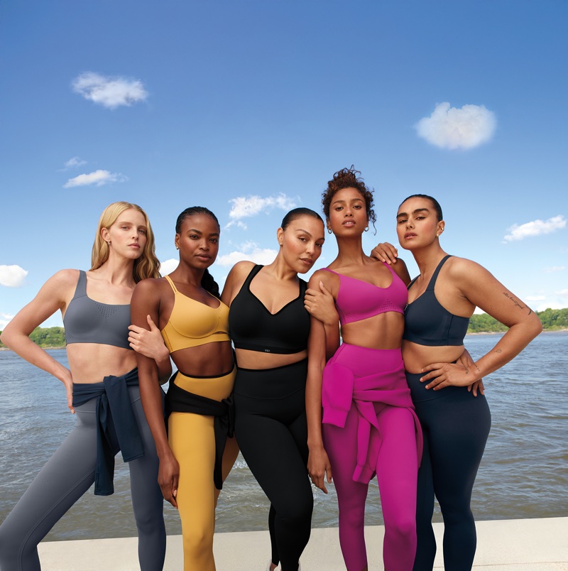 Five models standing side-by-side outdoors wearing colorful sports bras and leggings against water and blue sky.