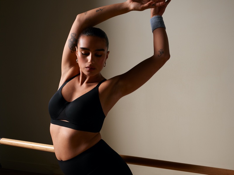 Model Jill Kortleve poses with arms overhead wearing a black sports bra and leggings beside a wooden barre in a studio setting.