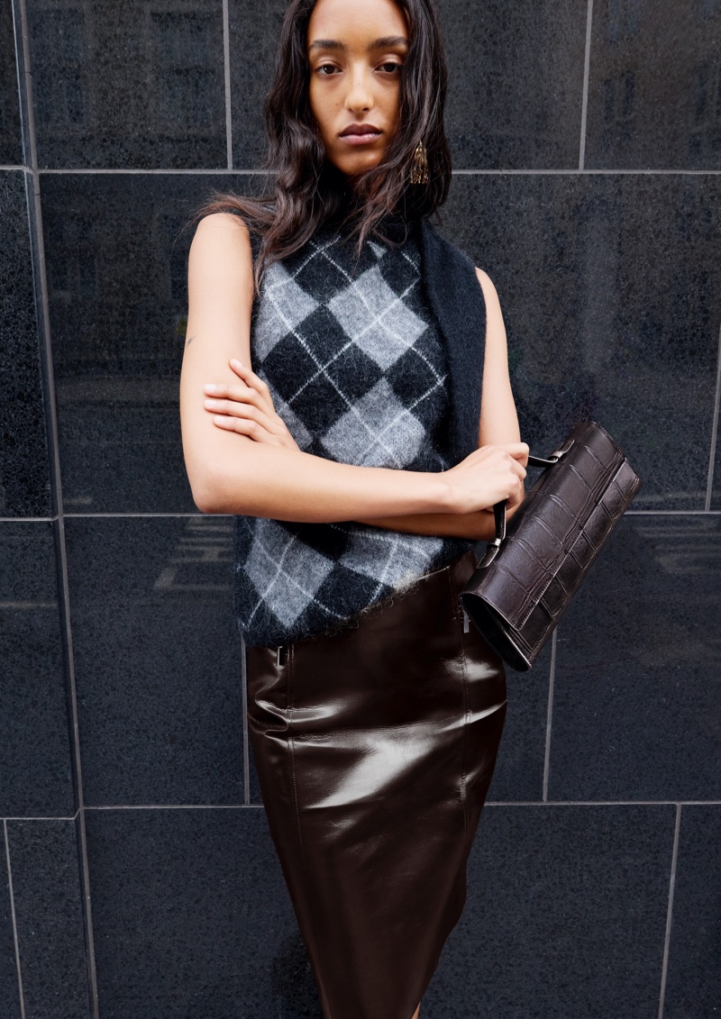 Model standing against a dark tiled wall wearing a grey argyle sweater vest, glossy brown skirt, and holding a textured handbag.