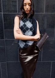 Model in argyle sleeveless sweater and dark leather skirt holding a brown handbag against a black stone wall