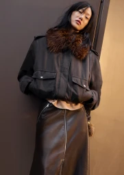 Sora Choi wearing a dark jacket with a fur collar and leather skirt leaning against a dark wall for H&M Fall-Winter 2025 campaign.