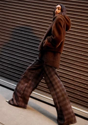 Model wearing a brown hooded fuzzy jacket and plaid wide-leg trousers standing against a slatted metal shutter backdrop.