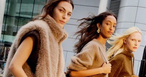 Three female models wearing brown textured autumn apparel outdoors against a modern glass and steel building exterior.