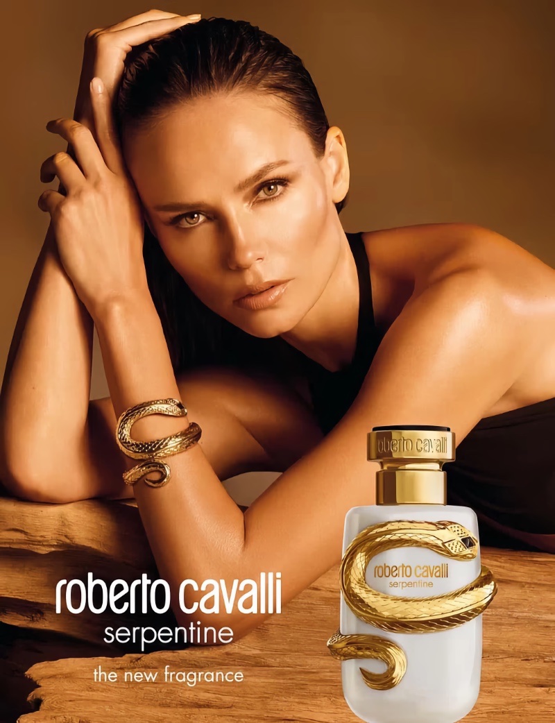 Model Natasha Poly posing with a Roberto Cavalli Serpentine fragrance bottle on a rustic wooden surface.