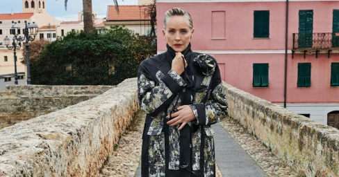Sharon Stone wearing a patterned black coat standing on a stone bridge with pink building background