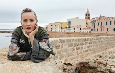 Sharon Stone resting her chin on her hand, wearing an olive patterned jacket outdoors near a stone wall and coastal town buildings.