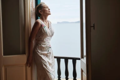 Sharon Stone wearing a white patterned dress leaning against an open doorway looking out toward the ocean.