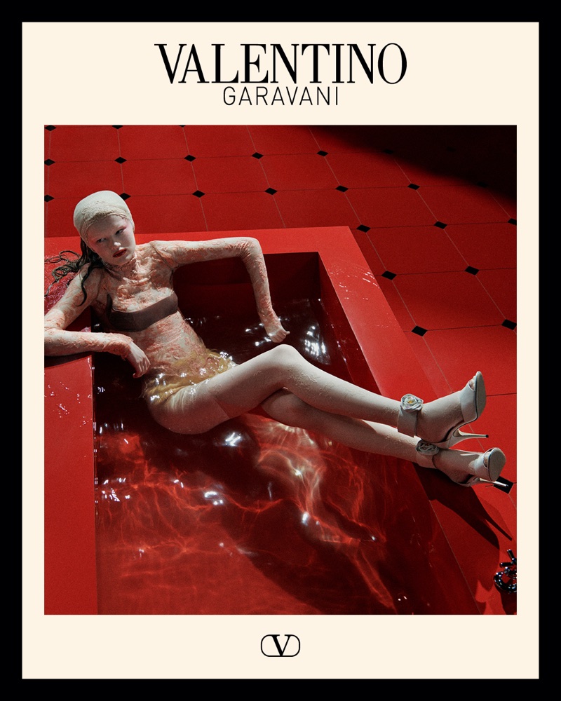 Model reclining in a shallow red tiled pool wearing high heels and headwrap in a Valentino Garavani fashion campaign.