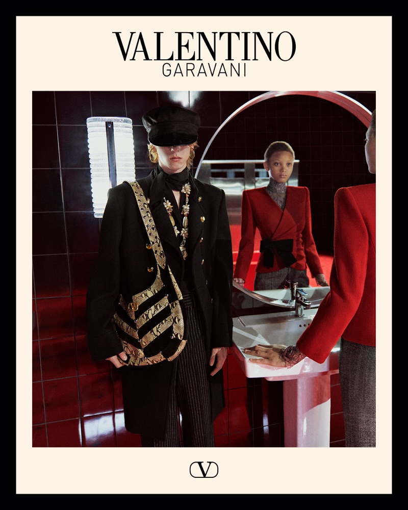 Model wearing a black coat, baker boy cap, and patterned shoulder bag standing beside a person in a red blazer in a tiled restroom.