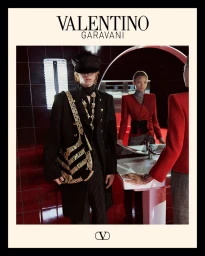 Clairo wearing a black coat and cap holding a patterned shoulder bag in a red tiled bathroom for Valentino shot by Glen Luchford.