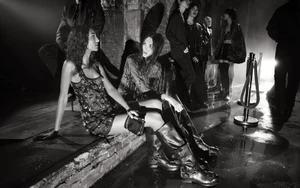 Two female models seated backstage in a dimly lit industrial space, wearing embellished clothing and tall leather boots