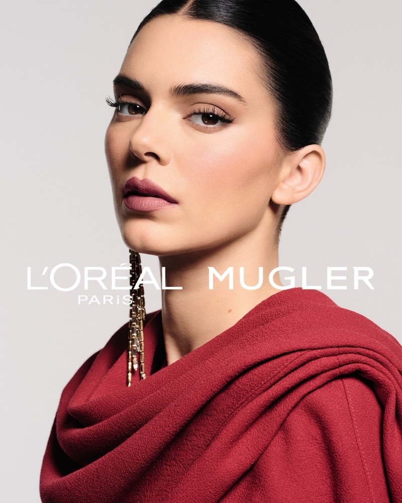 Kendall Jenner wearing a draped red cowl top and long gold earring against a pale studio backdrop with L'Oréal Mugler text.