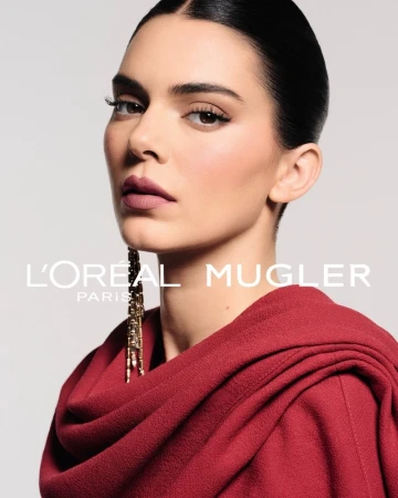 Kendall Jenner wearing a draped red cowl top and long gold earring against a pale studio backdrop with L'Oréal Mugler text.